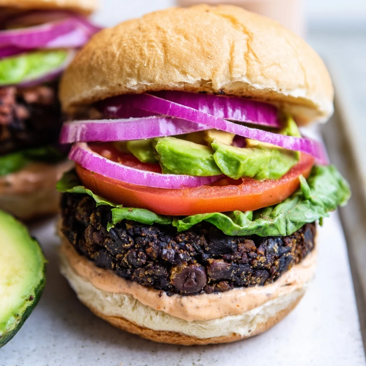 The Vegan Black Bean Burger with Chipotle Mayo sits on a rustic wooden board with sliced avocado and fresh lettuce leaves. 