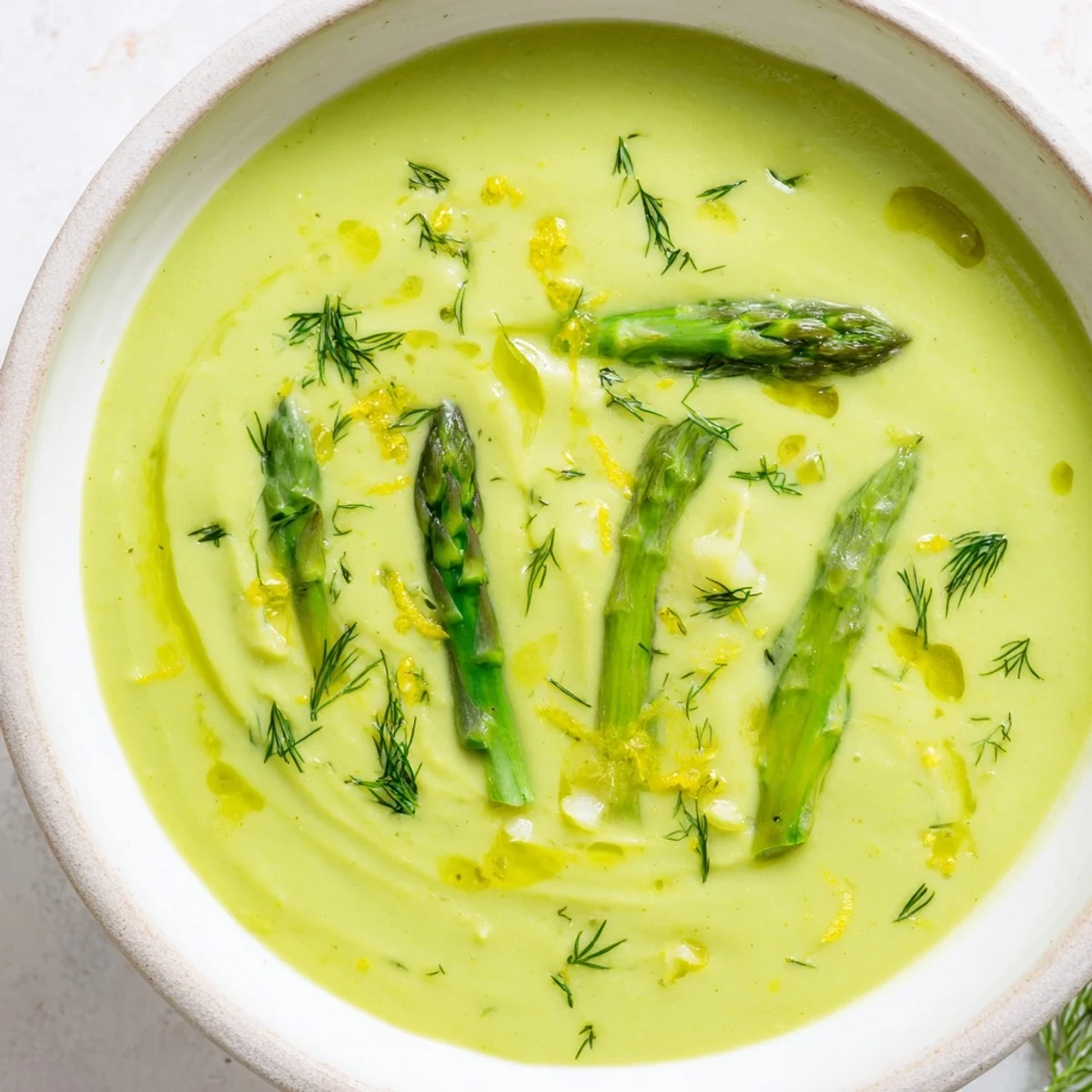 A close-up of creamy asparagus soup with lemon and dill garnish, served in a rustic white bowl.