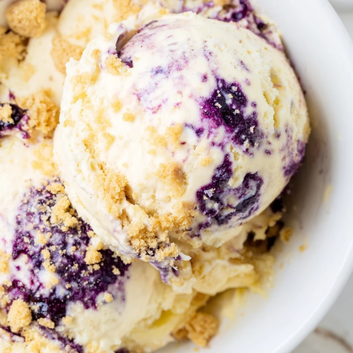 A scoop of homemade Lemon Blueberry Cheesecake Ice Cream with graham cracker crumbs in a chilled bowl.